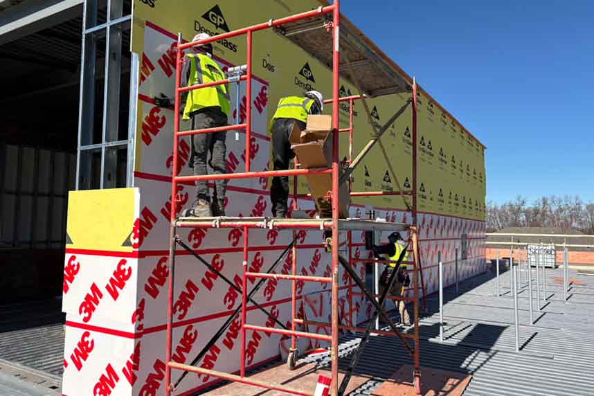 Cullen Insulation technicians on red scaffolding install 3M‑branded air‑barrier panels over green DensGlass® exterior sheathing on a steel‑framed commercial building under a clear blue sky.
