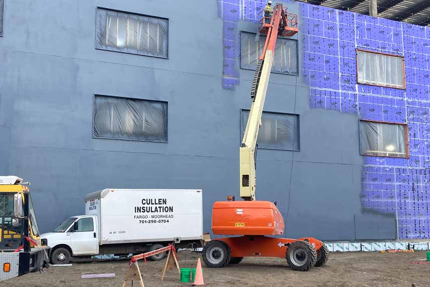 Boom‑lift installer spraying dark fluid air‑barrier coating onto purple sheathing of a multi‑story commercial building, with a Cullen Insulation trailer parked below.