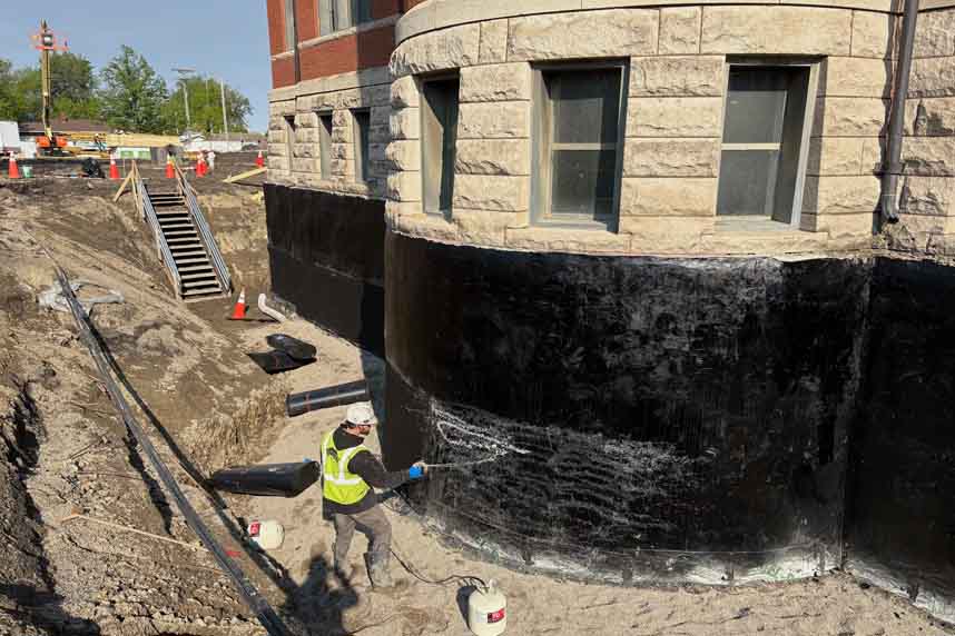 Worker rolling black waterproofing onto a curved stone foundation wall in an open excavation.