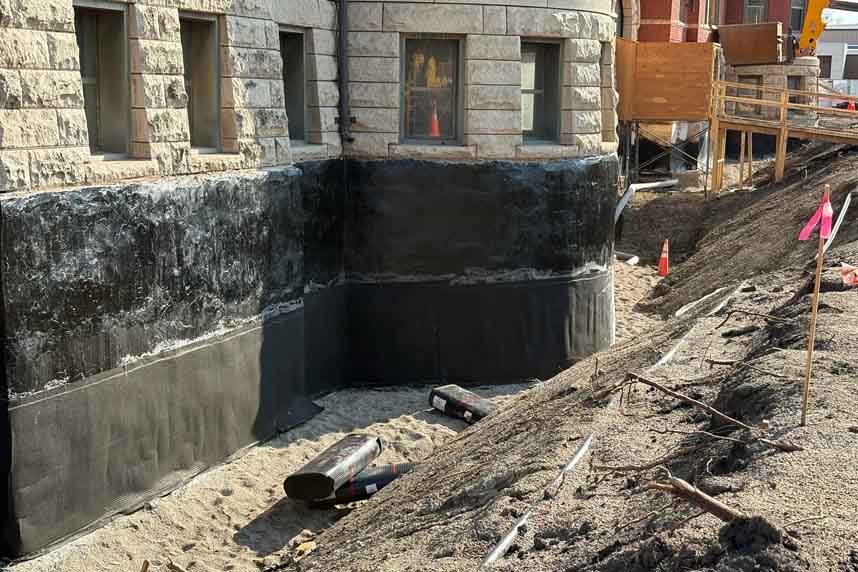 Dark membrane‑coated foundation corner with dimple board at the base and rolls of material nearby.