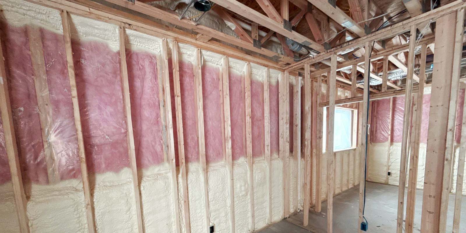 Interior view of a residential framing project with spray foam insulation on the lower half of the walls and pink fiberglass batt insulation on the upper half, showcasing a hybrid insulation method.