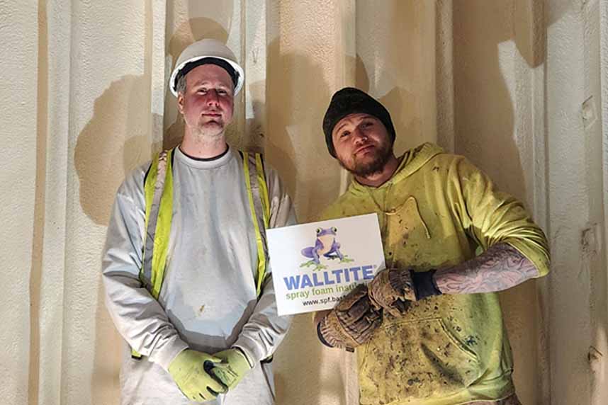 Two insulation installers standing in front of a wall covered with cured spray foam insulation holding a WALLTITE spray foam sign.