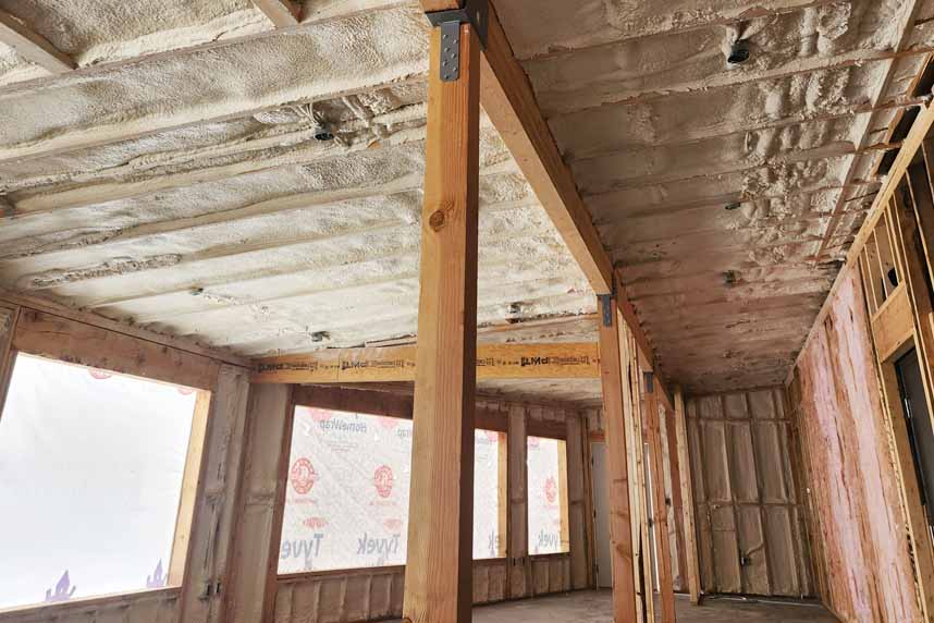 Framed living‑room under construction with white spray‑foam on ceiling and walls, Tyvek‑covered windows, and exposed studs.