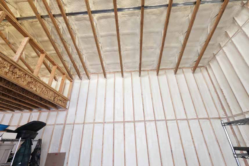 Tall walls and ceiling of a large room fully insulated with white spray foam between the wooden framing studs.