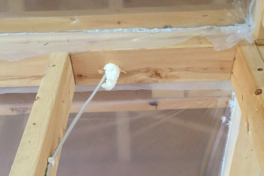 Clear plastic vapor‑barrier sheeting stapled to interior wall framing, with a small bead of spray foam sealing an electrical wire penetration through the stud.