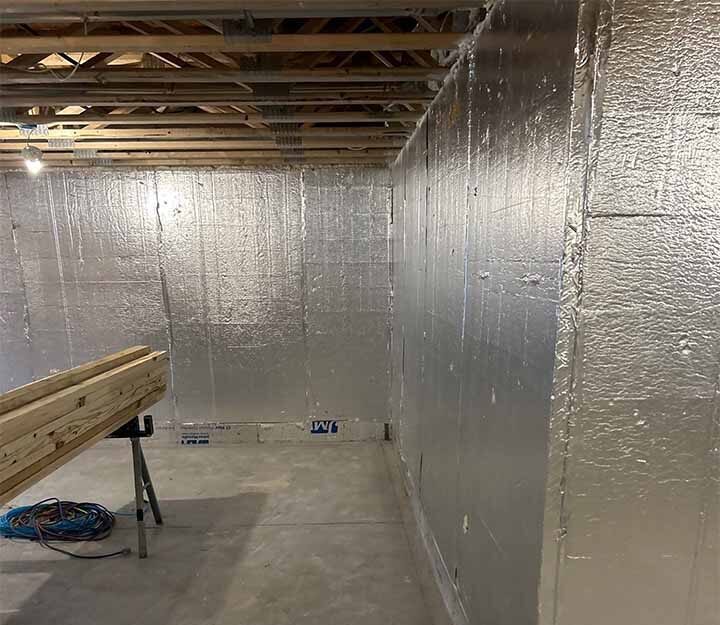 Silver‑faced rigid foam insulation panels cover basement walls behind exposed floor joists and construction materials.