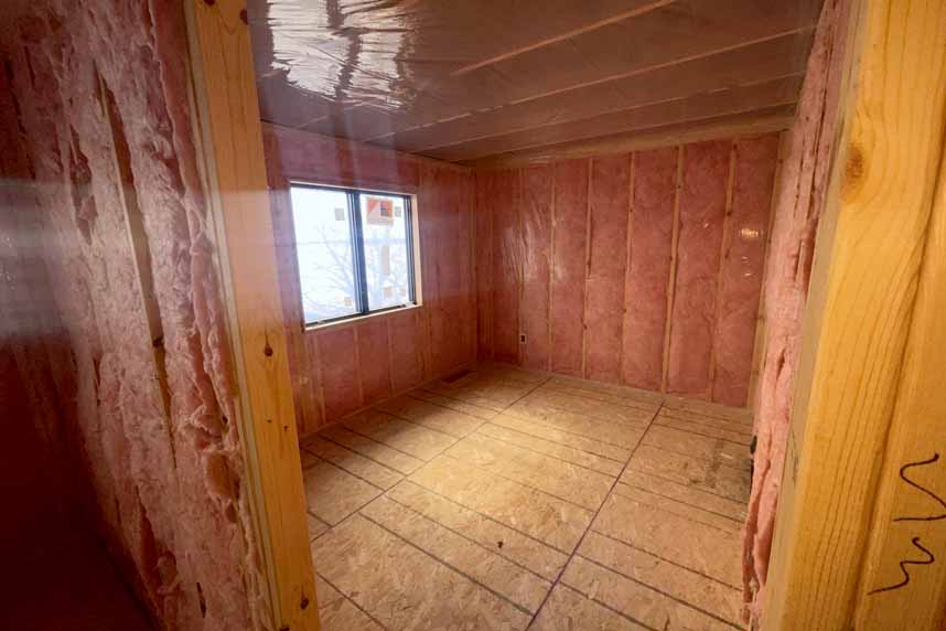 Interior view of a multifamily residential unit under construction showing framed walls with pink fiberglass insulation installed and an open window.