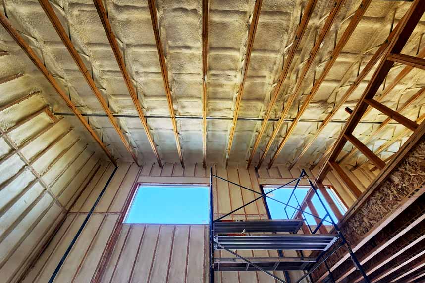 Interior view of a multifamily building under construction with spray foam insulation applied to walls and ceiling framing, showcasing an elevated work platform.