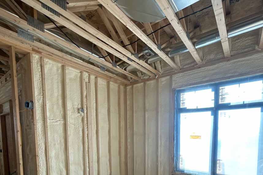 Residential interior showing newly installed spray foam insulation between wall studs and exposed mechanical ductwork overhead.