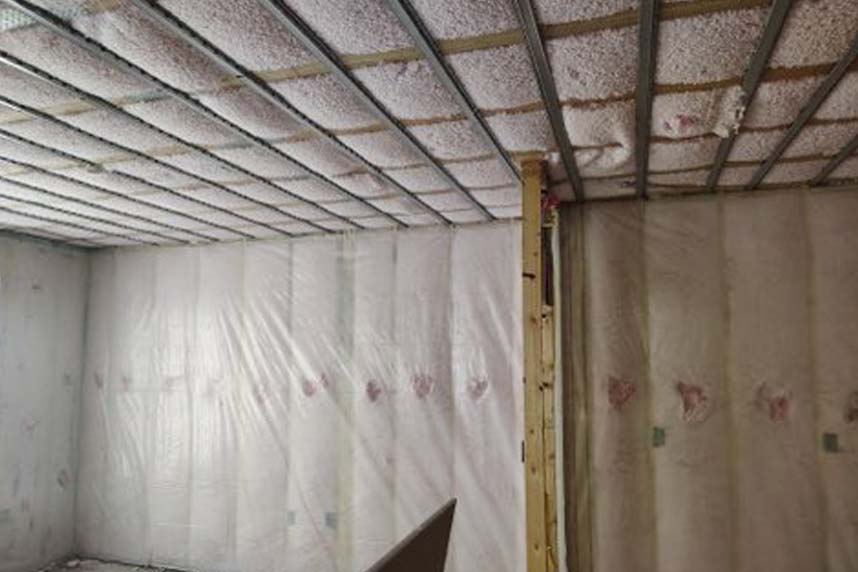 Interior room with unfaced fiberglass batts on ceiling, polyethylene‑covered wall batts and metal hat‑channel grid in multifamily build.