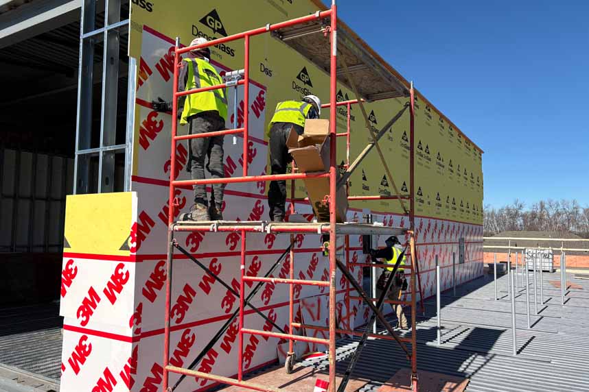 Two installers on scaffolding applying red‑logo 3M self‑adhered panels to a yellow DensGlass‑clad exterior wall at a commercial jobsite.